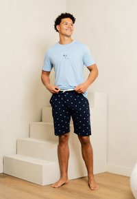 Světle modré bavlněné tričko s grafikou palmy. Tmavě modré šortky s bílým hvězdicovým vzorem a šňůrkovým pasem. Stojící na schodech.