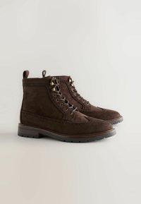 Bruine suède enkelboots met een wingtipt ontwerp, voorzien van messing oogjes en een gestructureerde zool. Inclusief een kleine trekflap voor gemakkelijke instap.
