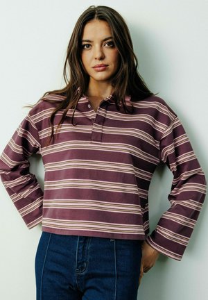 Jeune femme aux longs cheveux bruns portant un t-shirt rayé violet et rose à manches longues et un jean bleu, debout contre un mur blanc.