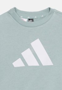 Ανοιχτό μπλε φούτερ με στρογγυλή λαιμόκοψη και μεγάλο λευκό λογότυπο Adidas στο κέντρο του στήθους.