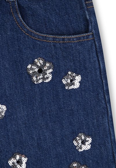 Tissu en denim de couleur bleu foncé, avec des motifs floraux en sequins argentés éparpillés. Comprend une poche soulignée par des surpiqûres contrastantes.