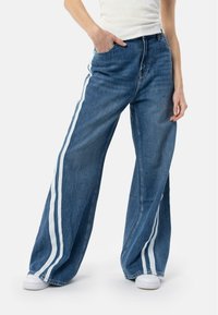 Šviesiai mėlynos plačios džinsinės kelnės su baltomis šoninėmis juostomis, aukštu liemeniu, priekinėmis kišenėmis ir švariai užbaigtais galiukais.