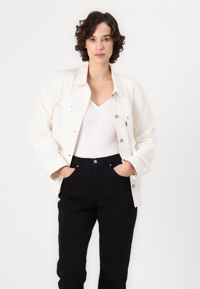 Chaqueta de mezclilla blanca con herrajes plateados y dos bolsillos, llevada sobre una blusa blanca y pantalones negros. Textura suave y corte relajado.