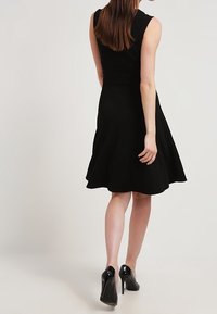 Schwarzes ärmelloses Kleid mit figurbetontem Oberteil und ausgestelltem Rock, aus glattem Stoff, kombiniert mit schwarzen High Heels.