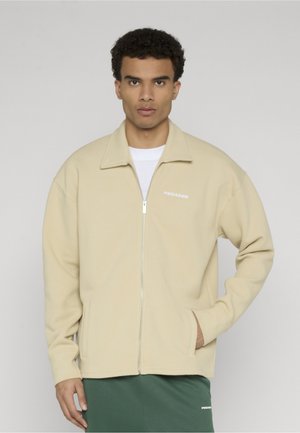 Mand iført beige jakke med lynlås over hvid skjorte og grønne bukser, stående med den ene hånd i jakkelommen, neutral baggrund.