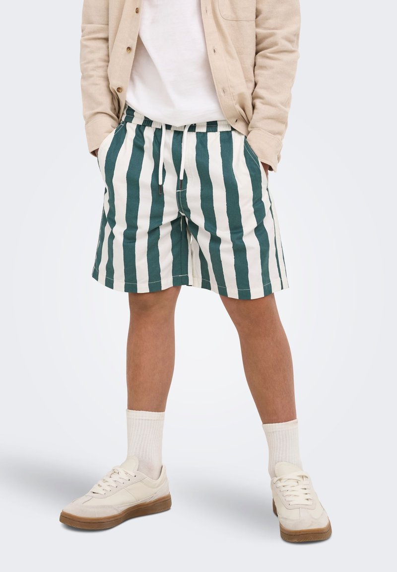 Homme portant un short rayé vertical vert et blanc, des baskets blanches, des chaussettes blanches, une veste beige et un t-shirt blanc, debout les mains dans les poches.