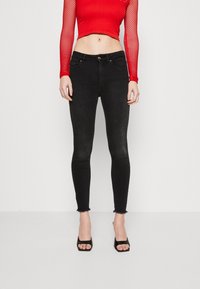 Mujer con top corto de malla de manga larga rojo, jeans pitillo negros de talle alto con dobladillos deshilachados y tacones abiertos negros.