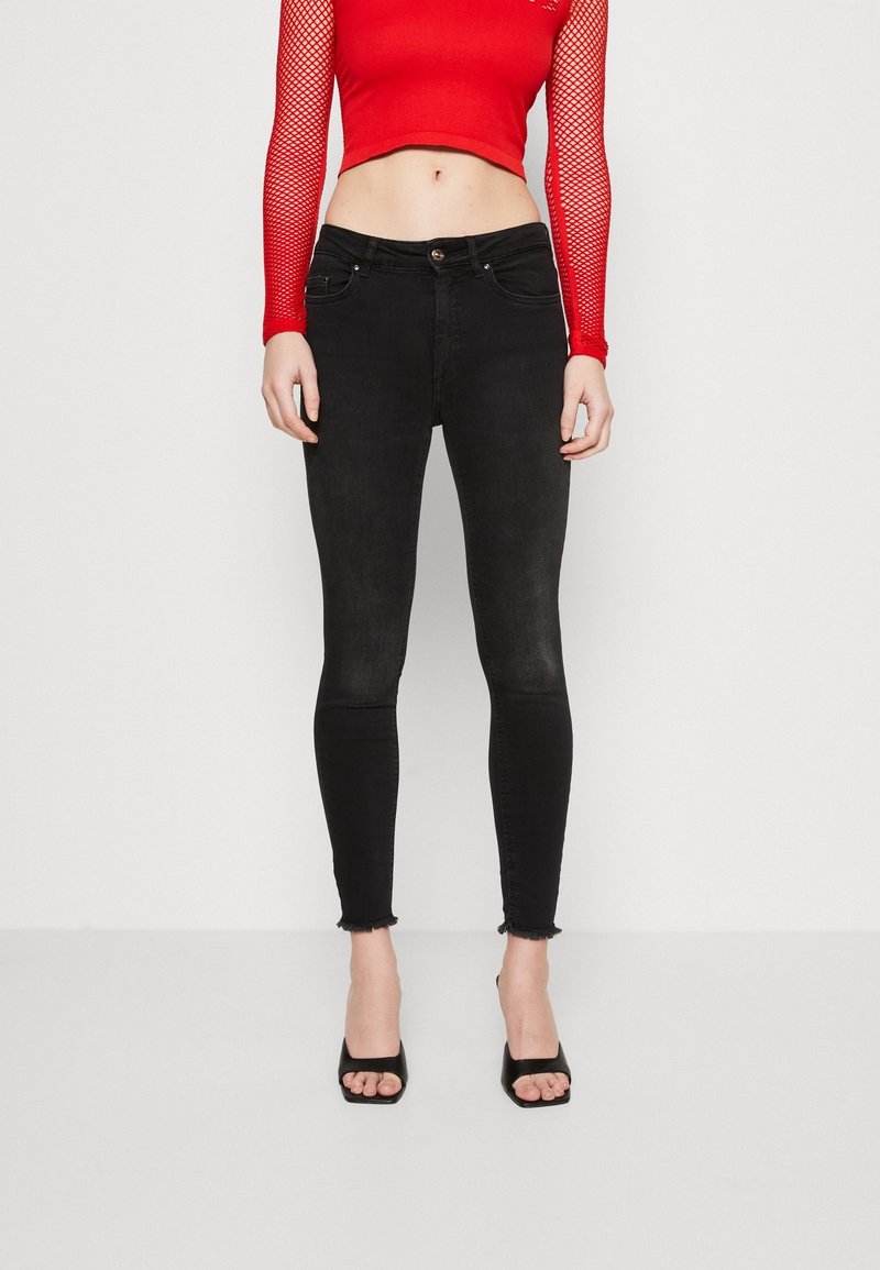 Mujer con top corto de malla de manga larga rojo, jeans pitillo negros de talle alto con dobladillos deshilachados y tacones abiertos negros.