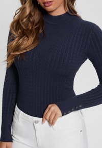 Marineblauer Rippstrickpullover mit hohem Kragen, der strukturierte Muster und Knopfdetails an den Ärmeln aufweist. Kombiniert mit weißen Hosen.
