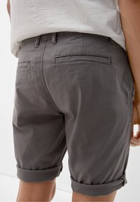Vue arrière d'une personne portant un short gris à ourlet roulé et une chemise blanche rentrée, se tenant devant un fond blanc.