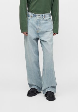 Person iført lyseblå jeans med vide ben, sorte chunky sko og en grøn langærmet skjorte, stående foran en ensfarvet baggrund.