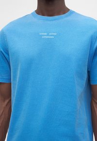 Homme portant un t-shirt bleu clair avec un texte blanc "SAMSØE SAMSØE COPENHAGEN" sur la poitrine sur un fond uni.
