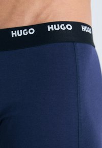 Marineblaue Boxershorts mit einem schwarzen elastischen Bund, der wiederholt mit weißen "HUGO"-Buchstaben bedruckt ist. Glatte Textur und körpernahe Passform.