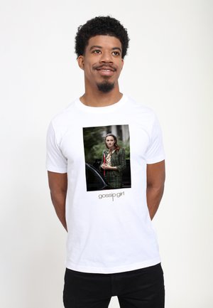 Mand iført en hvid T-shirt med et foto af en kvinde i en grøn ternet jakke, der holder en rød clutch, med teksten "gossip girl" nedenunder.