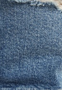 Närbild av blå denimtyg med synlig vävstruktur och fransig kant upptill.