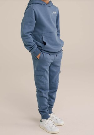 Kind draagt een blauw hoodie en bijpassende cargobroek van sweatstof met handen in de zakken, gecombineerd met witte sneakers op een beige achtergrond.