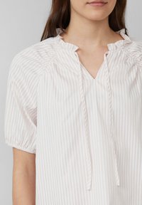 Biała bluzka z pastelowymi pionowymi paskami, z krótkimi bufiastymi rękawami, falbaniastym dekoltem i kołnierzykiem w kształcie litery V. Miękka, lekka tkanina.