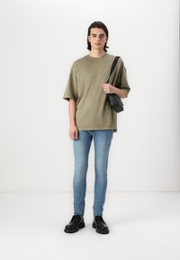 Løs olivengrøn t-shirt, lyseblå tætsiddende jeans, sort skuldertaske, sorte snørebåndssko, stående på en hvid baggrund.