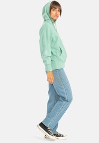 Mint green hoodie with a front pocket and cuffed sleeves, featuring a graphic detail on the left sleeve, paired with loose blue jeans and black sneakers.