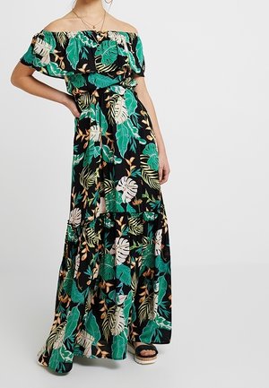 Mujer con un vestido largo negro sin hombros con estampado de hojas tropicales verdes y beige, de pie con una mano en la cadera y llevando sandalias de plataforma.