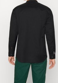 Camisa negra de manga larga con cuello puntiagudo, puños abotonados y dobladillo ligeramente curvado. La tela parece lisa, con detalles de costura sutiles.