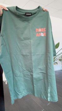 Camiseta de algodón verde con texto impreso "MORE AMOR" en rosa, con acentos florales. Mangas cortas y escote redondo. Textura suave.