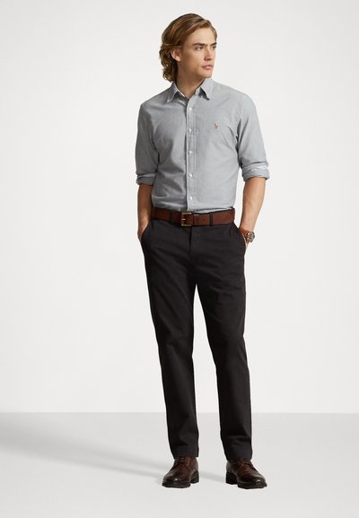 Chemise bleu clair à manches retroussées, pantalon noir, ceinture en cuir marron, et chaussures marron. La chemise comporte un petit logo sur la poitrine.