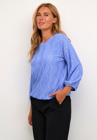 Lichtblauwe blouse met golfpatroon oogjes ontwerp, ronde halslijn en driekwartmouwen. Gecombineerd met zwarte broeken.