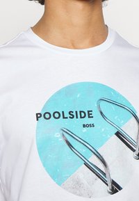 Bílý tričko s krátkým rukávem, které obsahuje kruhovou grafiku s tyrkysovou vodou a bazénovým žebříkem. Text říká "POOLSIDE BOSS" tučným písmem.