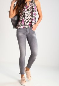 Blusa con motivo floreale e volant, abbinata a jeans grigi skinny e sneakers rosa pallido. Borsa nera visibile su uno sfondo chiaro.