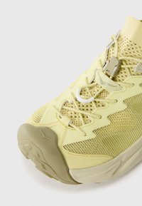 Chaussure de sport jaune avec une tige en mesh et un tissu texturé, un système de laçage visible et une semelle rembourrée avec des motifs de traction flexibles.