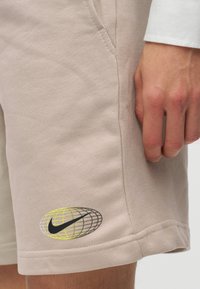 Κοντινό πλάνο μπεζ σορτς με ένα μαύρο και κίτρινο λογότυπο Nike σε σχήμα γης κοντά στο τελείωμα και ένα χέρι να ακουμπάει δίπλα στην τσέπη.