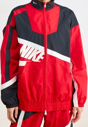 Osoba nosząca czerwoną i czarną kurtkę sportową Nike z białymi ukośnymi paskami oraz pasujące czerwone spodnie dresowe.