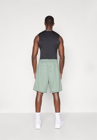 Schwarzes ärmelloses Sportoberteil und hellgrüne Shorts mit elastischem Bund, getragen mit weißen Socken und Schuhen, von hinten betrachtet.