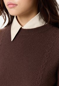 Kvinna som bär en brun texturerad tröja över en beige skjorta med krage, synlig från nacken till nedre delen av ansiktet.