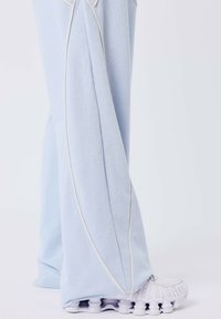 Lichtblauwe broek met witte piping, gedragen over witte sneakers met een kenmerkende gesegmenteerde zool, tegen een egale achtergrond.