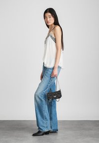 Femeie cu păr lung și închis la culoare, purtând un top fără mâneci de culoare albă și blugi largi de culoare albastră, ținând o geantă mică din material matlasat de culoare neagră cu curea din lanț.