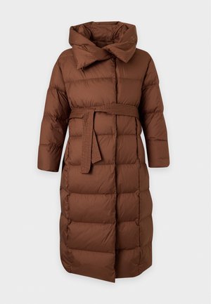 Une femme présente un long manteau matelassé marron par-dessus un pull beige et un pantalon noir, sur un fond blanc uni.