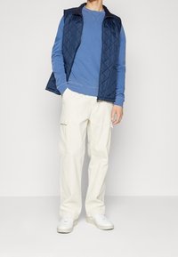 Marineblauer gesteppter Weste über einem blauen Sweatshirt, kombiniert mit cremefarbenen Cargo-Hosen und weißen Turnschuhen. Das Outfit hat eine lockere Passform und einen geschichteten Look.