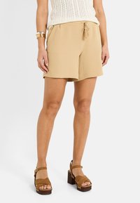 Pantalones cortos beige de corte relajado con cintura elástica y cordón ajustable. La textura es ligeramente arrugada. Combinado con sandalias de plataforma marrones.