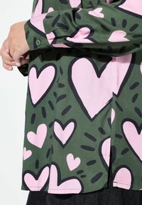 Groene shirt met een patroon van roze harten omrand door zwart. Lange mouwen met knoopmanchetten en zijsplitten aan de zoom.