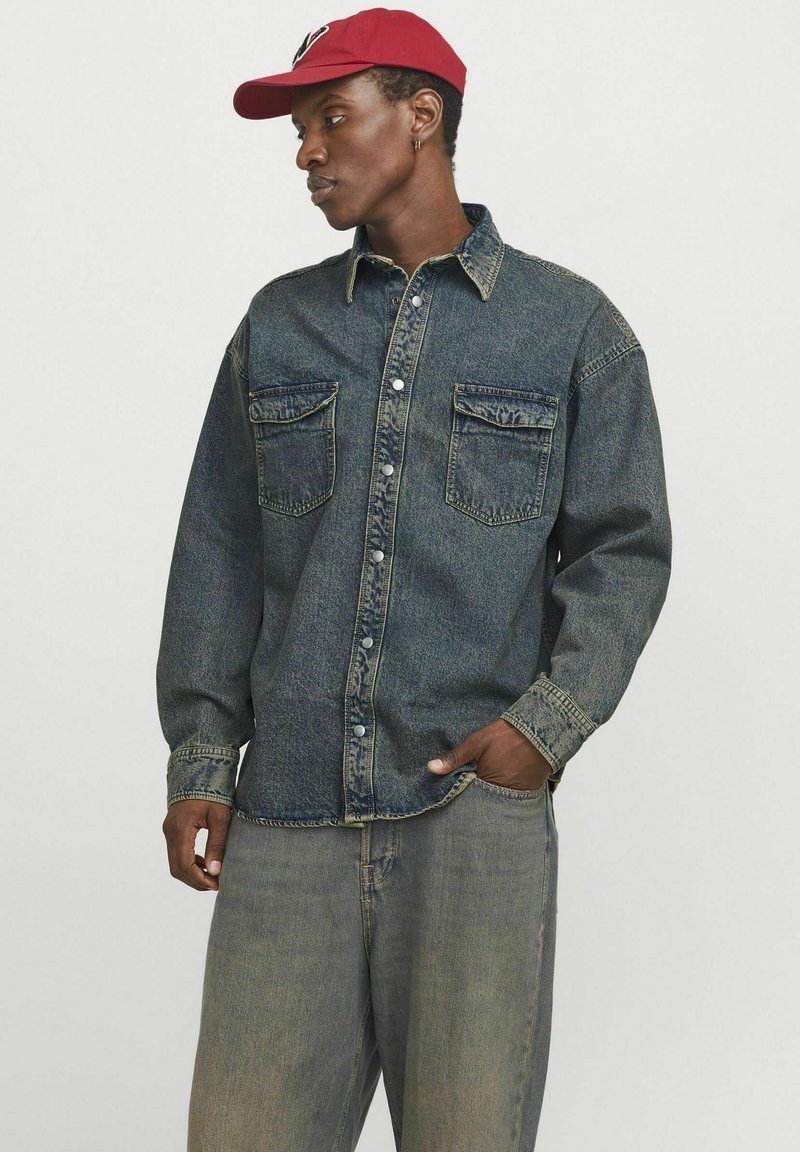 Camicia di jeans con bottoni anteriori, due tasche frontali e maniche lunghe. Indossata con pantaloni in denim chiaro e un berretto rosso.