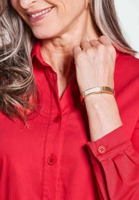 Rotes Hemd mit Knöpfen und glatter Textur, mit einem Umlegekragen am Ärmel. Ein dünnes goldenes Armband mit rechteckigem Design am Handgelenk.