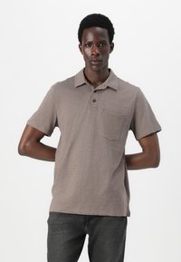 Camisa polo de manga corta, color marrón claro, con cuello y tres botones, y un bolsillo en el pecho. Material de algodón con un acabado texturizado.