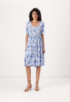 Sininen ja valkoinen kukkakuosiset mekko, jossa on lyhyet puffit, neliömäinen pääntie ja kerroksellinen hame. Yhdistetty hartstrapilla varustettujen vaaleanbeigejen kenkien kanssa.