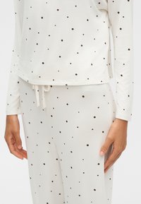 Gros plan d'une personne portant un haut blanc à manches longues et un pantalon à petits pois noirs avec une taille à cordon.