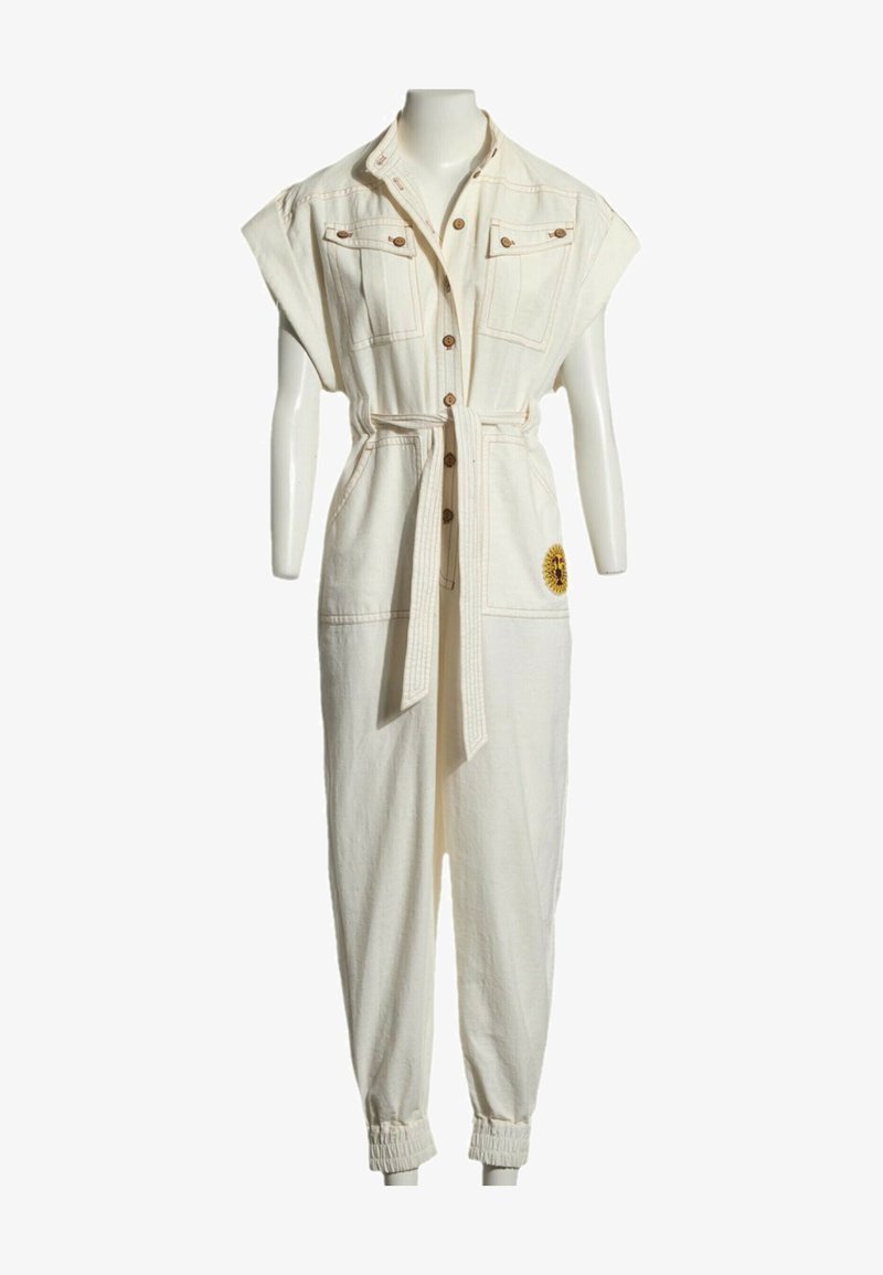 Combinaison sans manches couleur crème avec boutonnière sur le devant, ceinture à la taille, poches poitrine, poignets élastiques et patch tournesol sur la jambe droite.