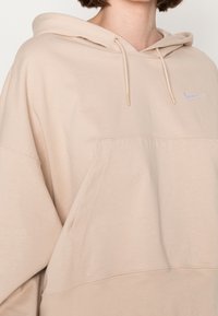 Beige oversized hoodie made of soft fabric, featuring a front pocket, drawstring hood, and a small blue Nike logo on the chest.