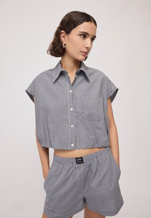 Junge Frau mit kurzen welligen Haaren trägt ein schwarz-weiß gestreiftes, bauchfreies Shirt und passende karierte Shorts, steht mit den Händen in den Taschen.
