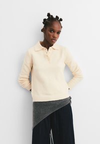 Tânără cu coafură în noduri bantu, purtând un pulover crem cu nasturi și o wrap asimetrică gri peste pantaloni black largi, stând în fața unui perete alb.